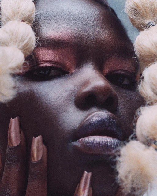 Close-up portrait of a woman with dark skin, copper metallic nails and makeup, and blonde braided hair in a beauty and fashion aesthetic.