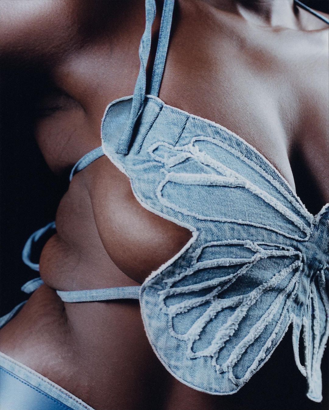 Close-up of an adult female torso wearing a light blue denim butterfly-shaped bikini top against a dark background in studio lighting.