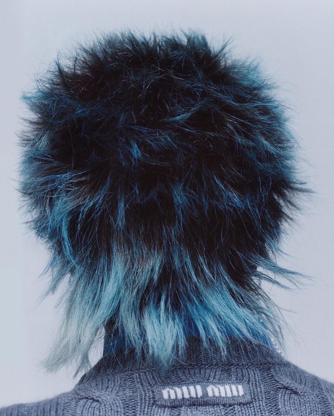 Close-up from behind of a person with a black and blue mullet, wearing a gray sweater with the 'Miu Miu' logo at the neckline.