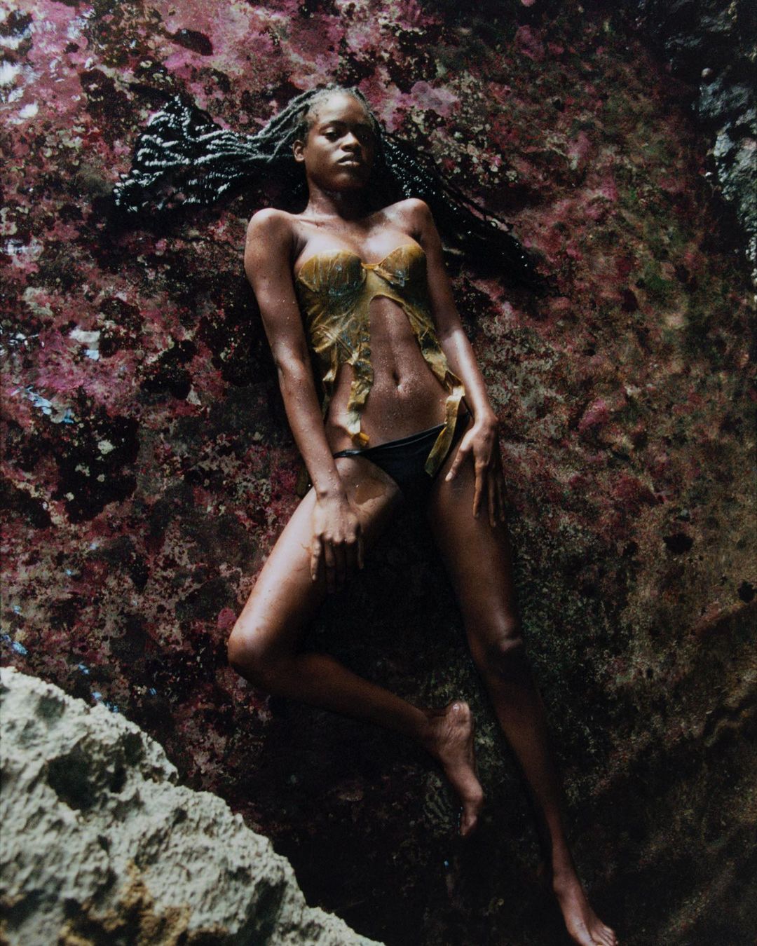 Woman with dark braided hair, wearing a leaf bra and bikini bottoms, lies on rock in an outdoor setting near the ocean.