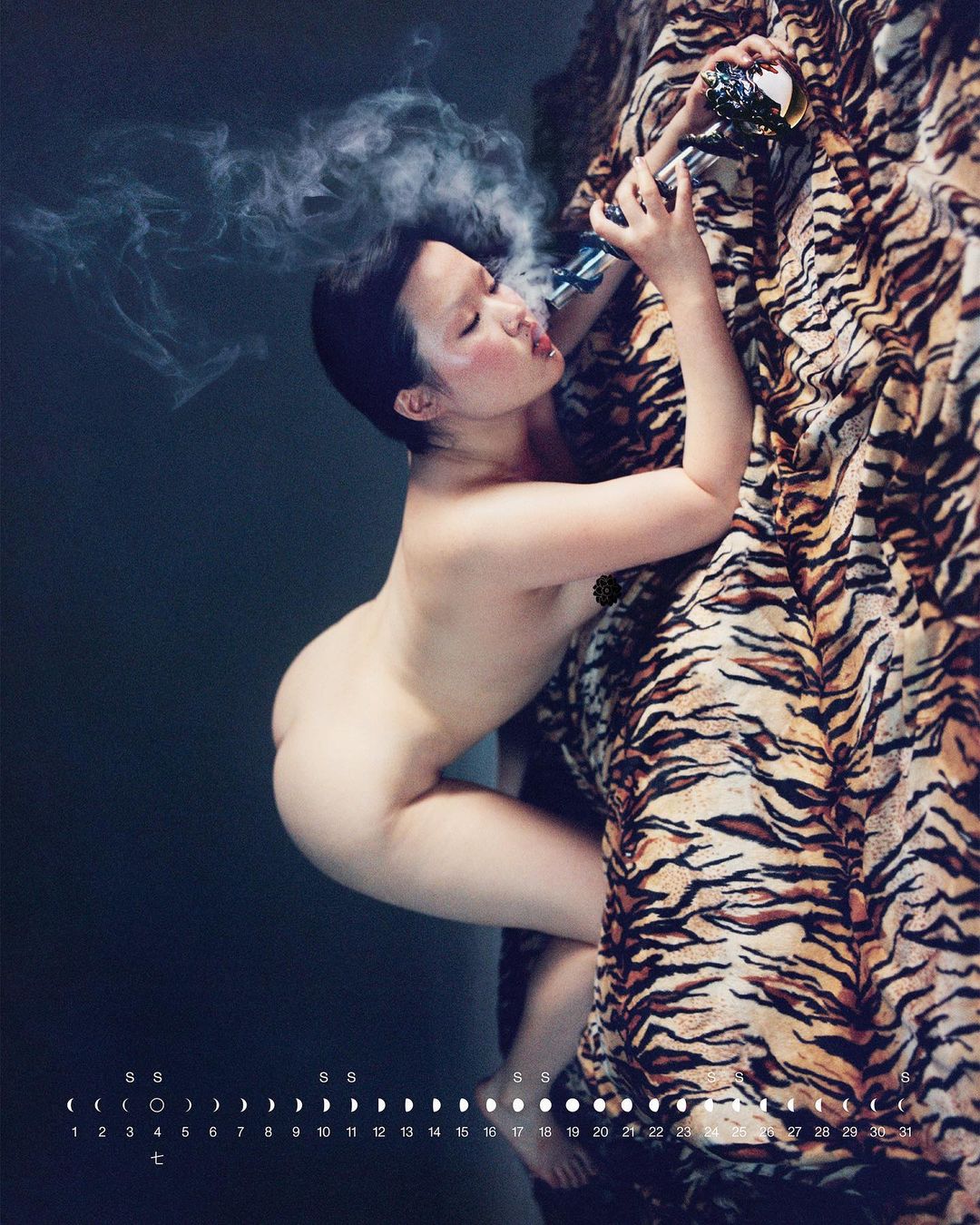 Nude adult female is smoking from a glass pipe, posing next to a tiger-print drape in a photo studio with a dark blue wall background.