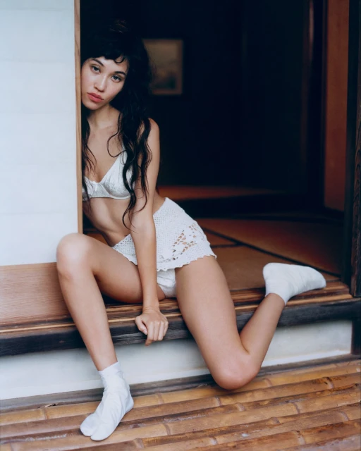 A fair-skinned woman with dark hair poses inside a Japanese-style room, wearing white crochet lingerie and socks, seated on a tatami floor.