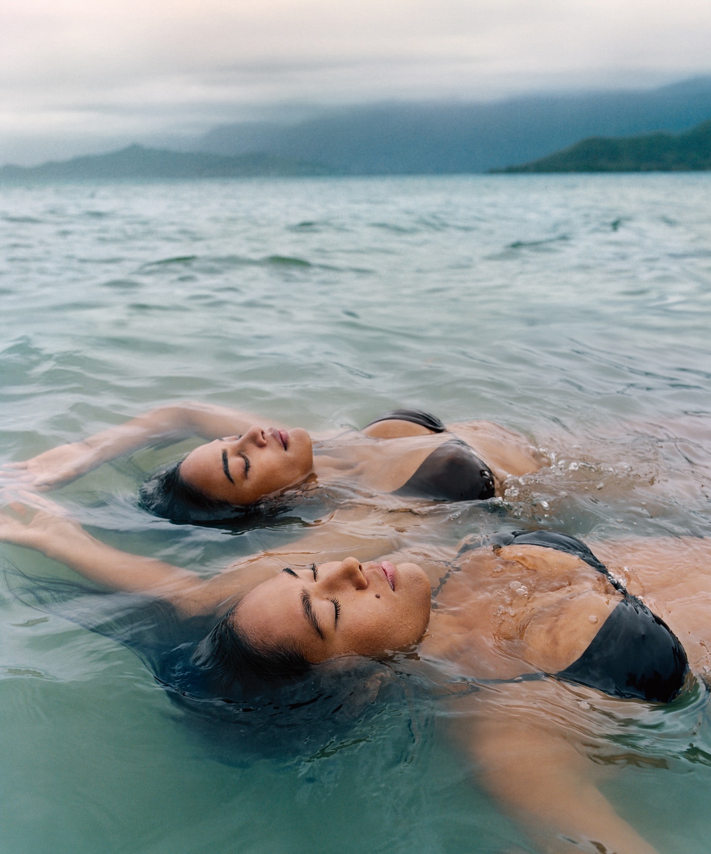 Two adult women in black bikini tops float in the ocean, eyes closed, with a distant island and cloudy sky visible in the background.