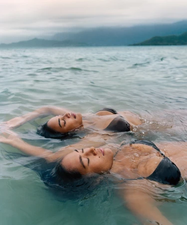 Two adult women in black bikini tops float in the ocean, eyes closed, with a distant island and cloudy sky visible in the background.