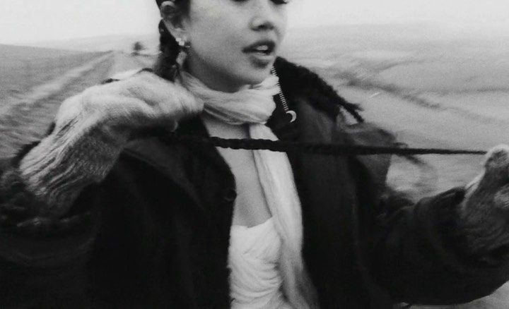 Monochrome close-up of a woman in a coat and scarf standing in a field, with braids, earrings, and a somber expression.