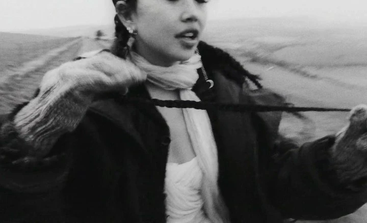 Monochrome close-up of a woman in a coat and scarf standing in a field, with braids, earrings, and a somber expression.