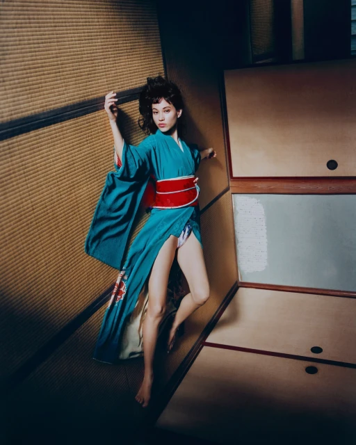 A woman in a teal kimono with a red obi lies on a tatami mat, with light brown paneling in the background, in a low-angle medium shot.