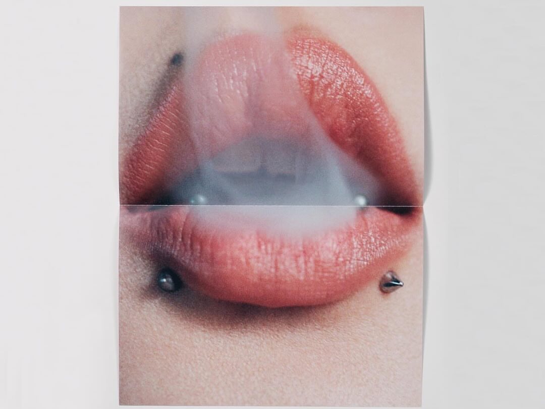 Close-up of a woman's mouth with pink lipstick and lip piercings exhaling smoke. The image is divided into two sections by a fold.