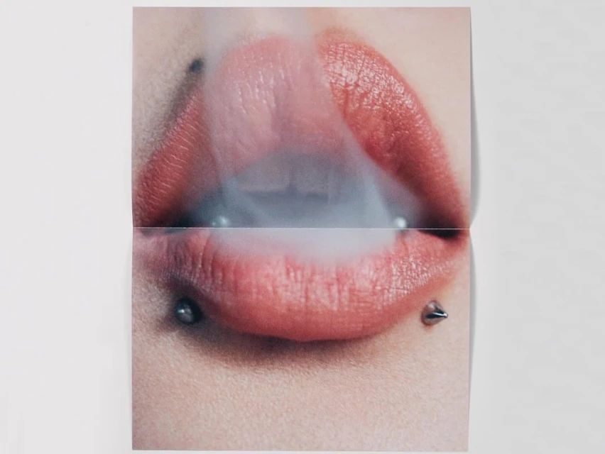 Close-up of a woman's mouth with pink lipstick and lip piercings exhaling smoke. The image is divided into two sections by a fold.