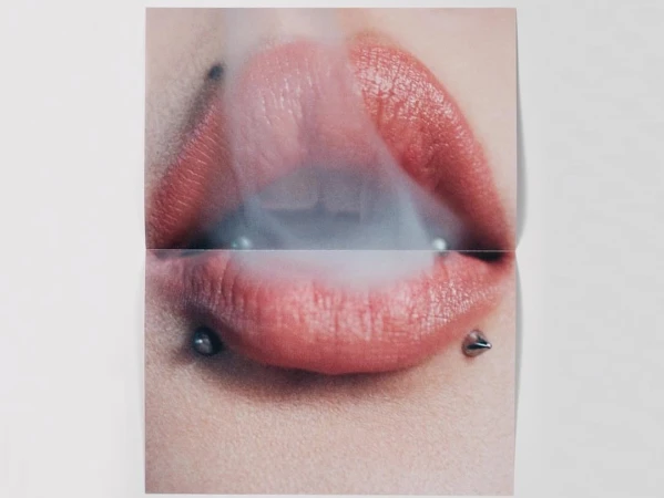Close-up of a woman's mouth with pink lipstick and lip piercings exhaling smoke. The image is divided into two sections by a fold.