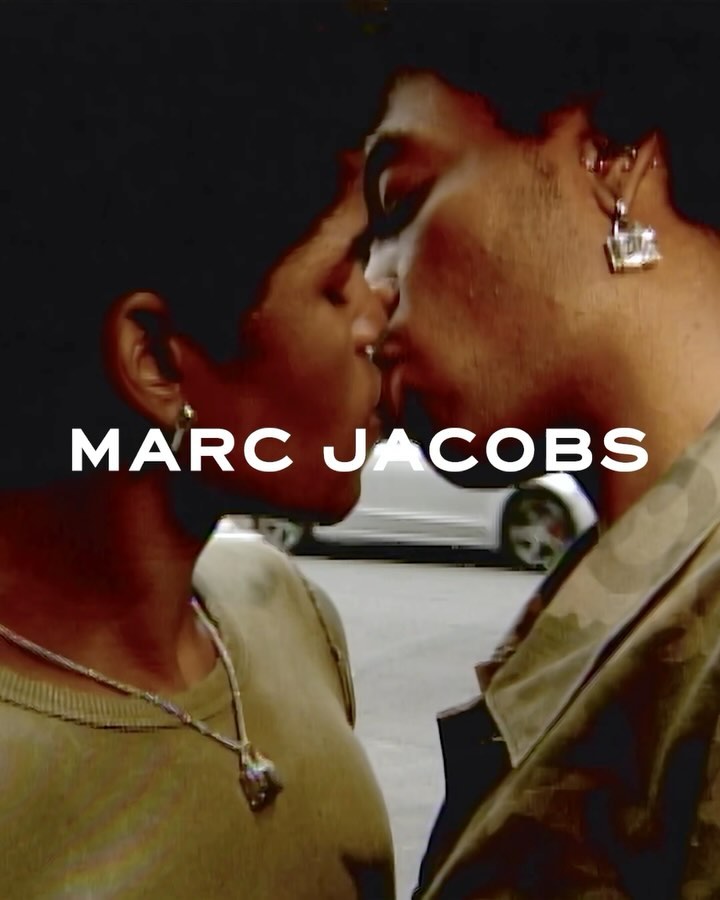 Close-up of two adults kissing with gold jewelry, daytime street background and the brand logo, MARC JACOBS, in white letters.