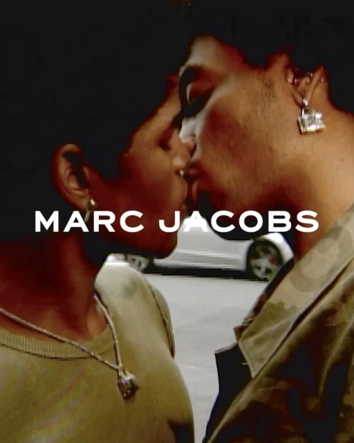 Close-up of two adults kissing with gold jewelry, daytime street background and the brand logo, MARC JACOBS, in white letters.