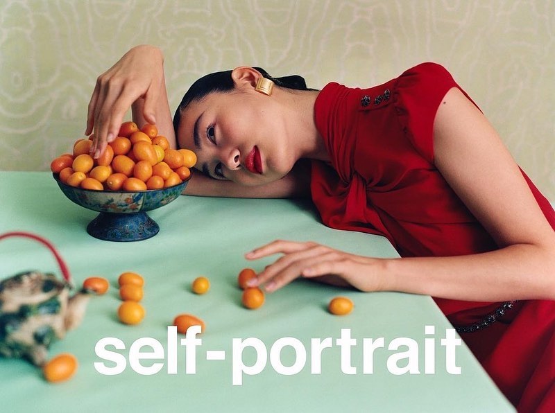 Portrait of an Asian woman in a red dress leaning on a table with kumquats in a bowl and scattered on the table, indoor setting.