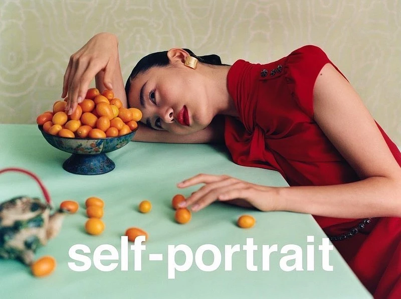 Portrait of an Asian woman in a red dress leaning on a table with kumquats in a bowl and scattered on the table, indoor setting.