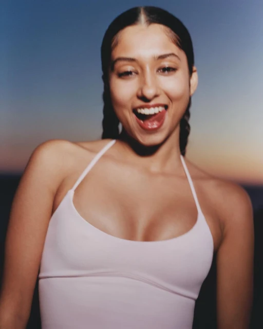 Close-up portrait of a smiling young woman with braided hair, wearing a light pink top, against a sunset background with a blue sky.