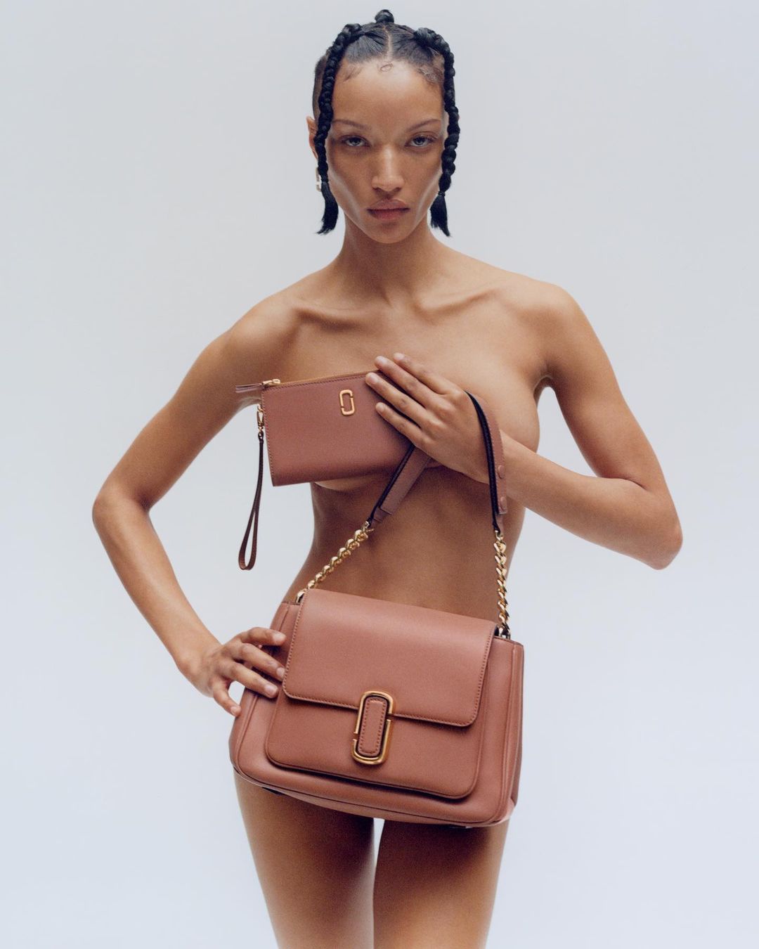 A nude-presenting adult woman with braids, holding a brown leather Marc Jacobs shoulder bag and matching wallet against a white background.