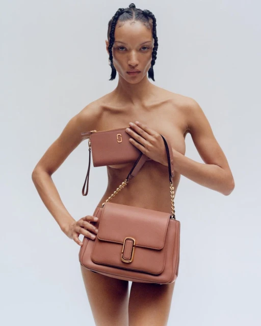 A nude-presenting adult woman with braids, holding a brown leather Marc Jacobs shoulder bag and matching wallet against a white background.