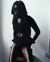 Portrait of a black woman with braided hair wearing a black top and belt seated against a white wall.