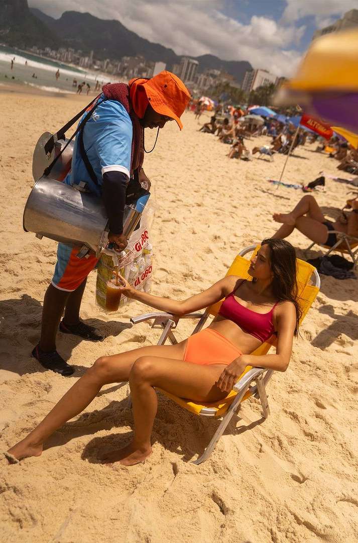 A vendor pours a drink for a woman on a beach. She is sitting in a beach chair and wearing a red top and orange bottoms.