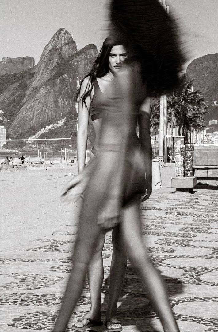 Monochrome portrait of a woman in swimwear with a blurred figure walking on a patterned path, mountains and palm trees in the background.