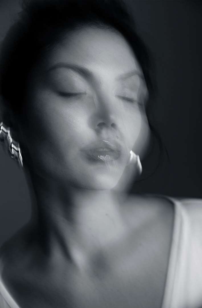 Monochromatic portrait of an adult woman with closed eyes and glossy lips, wearing statement earrings and a white top, with soft lighting.