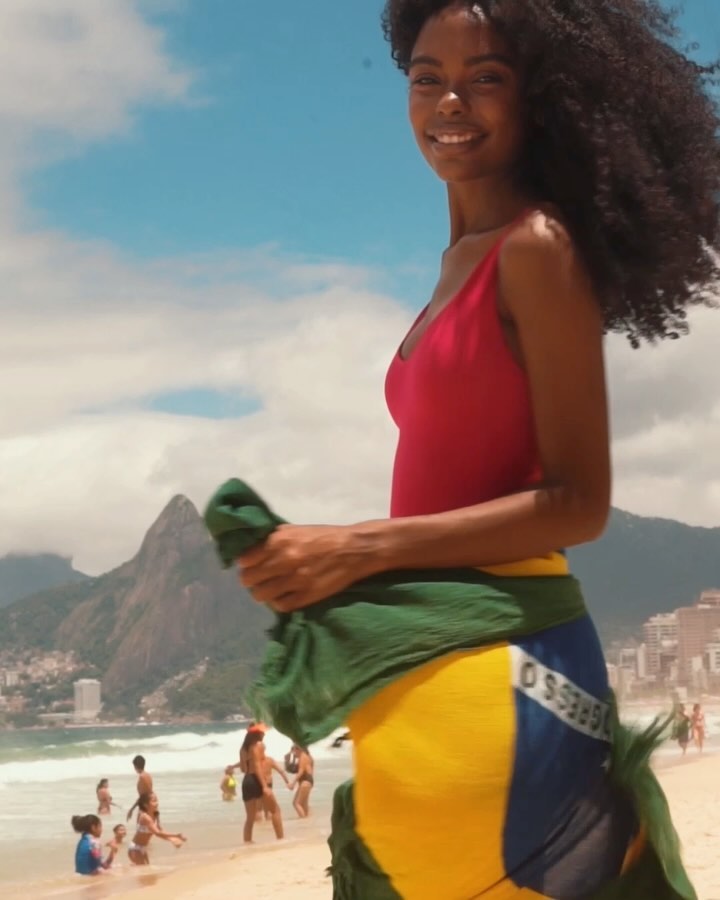 A smiling woman in a red swimsuit stands on a sandy beach with mountains and people in the background, wearing a Brazil flag sarong.