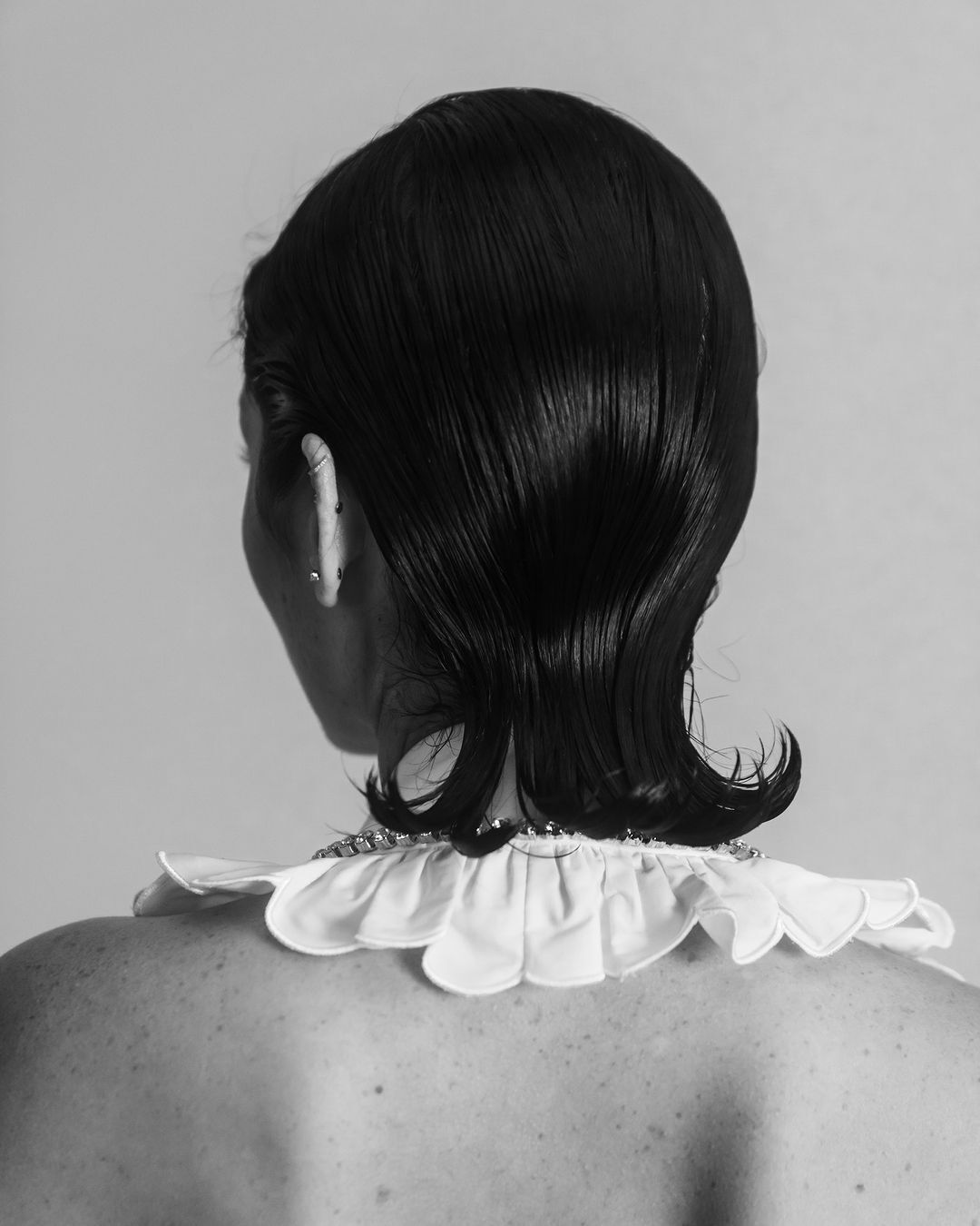 Back view of a person with short, styled dark hair, white ruffled collar, and exposed back, set against a bright, neutral background.