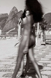 Black and white portrait of a woman in swimwear with motion blur on a beach walkway with mountain peaks in the background.