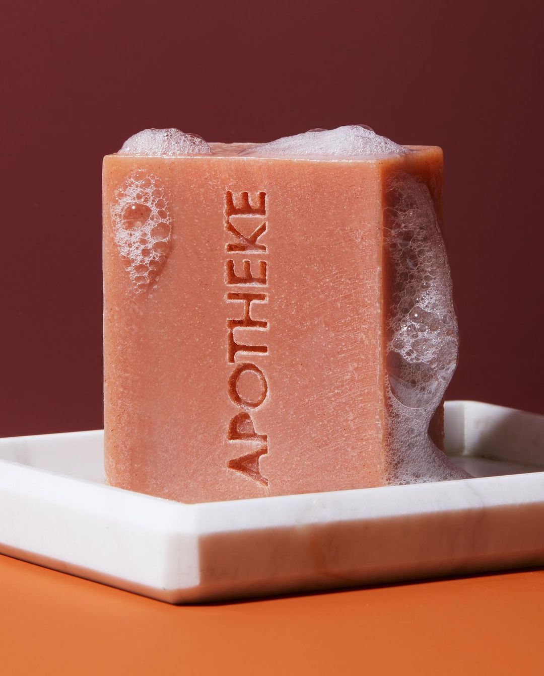 Close-up of a square terracotta soap bar on a white marble tray, with visible soap bubbles on a terracotta and burgundy background.