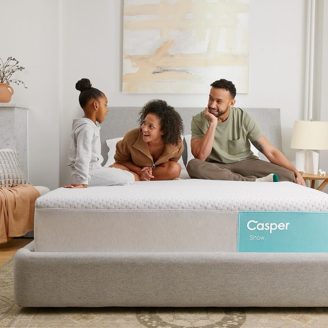 Family of three smiles and sits on a Casper Snow mattress in a brightly lit bedroom with off-white walls and neutral decor.