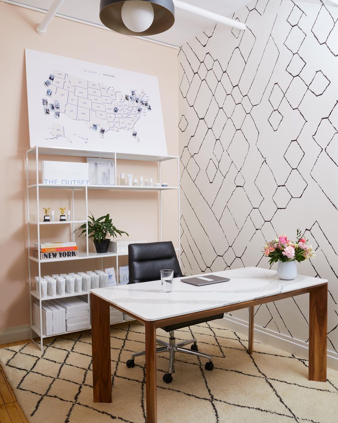 Modern office setup featuring a white marble-top desk with wood legs, a black leather chair, white shelving, and a beige diamond-patterned rug.