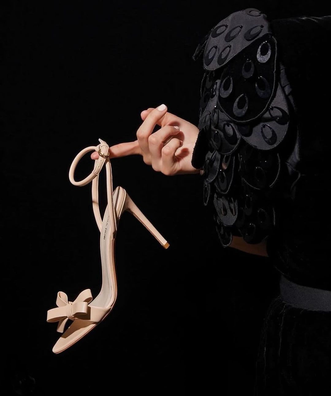 A woman's hand delicately holds a beige high-heeled sandal against a stark black background, creating a minimalist fashion image.