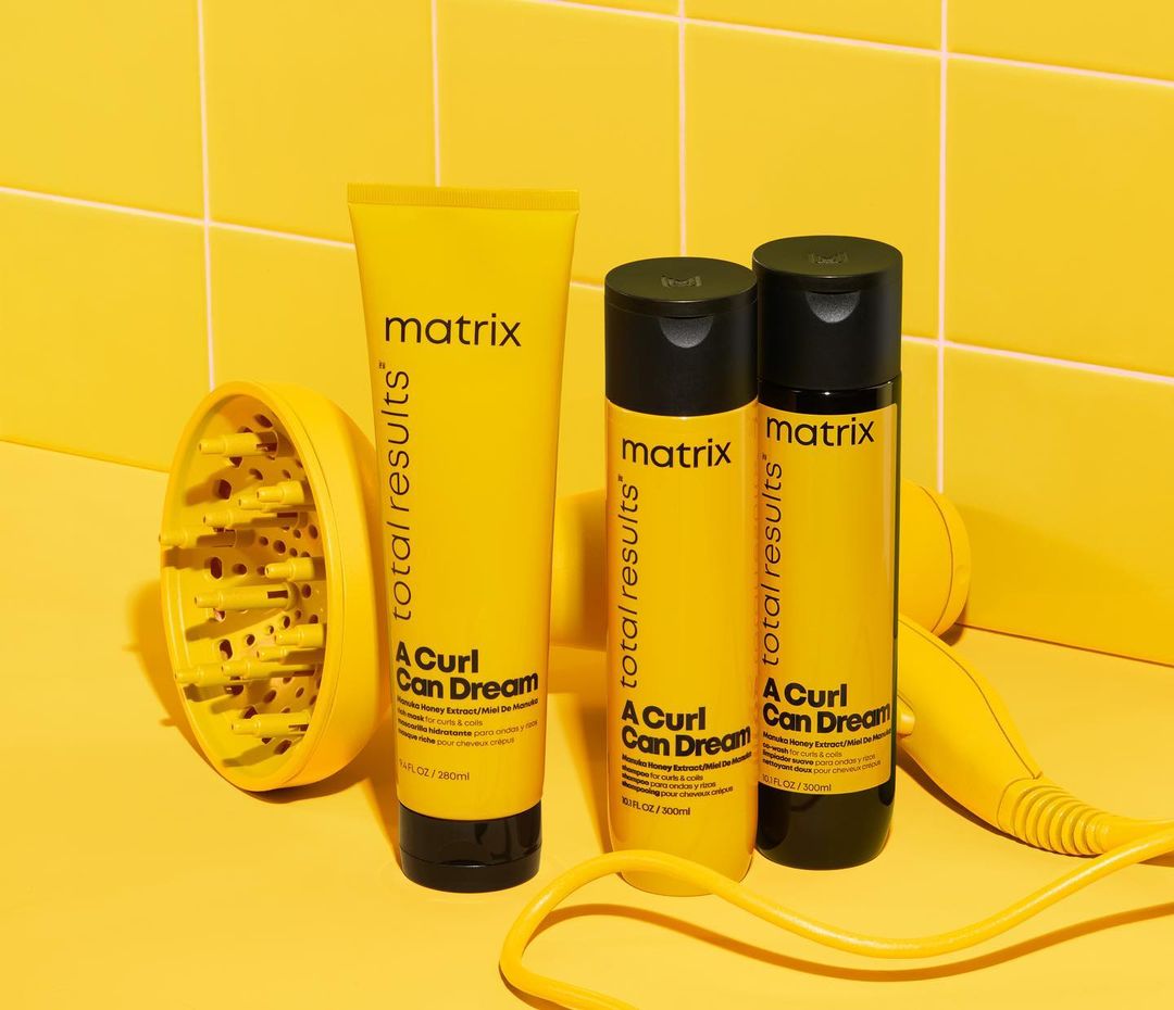 Matrix Total Results A Curl Can Dream product line in yellow packaging, including shampoo, conditioner, and diffuser, against a tiled yellow wall.