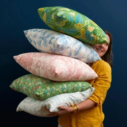 Woman in a yellow shirt holding a towering stack of patterned decorative throw pillows against a dark blue wall.