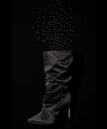 Black slouchy stiletto boot on a dark background with scattered small white dots floating upwards from the top.