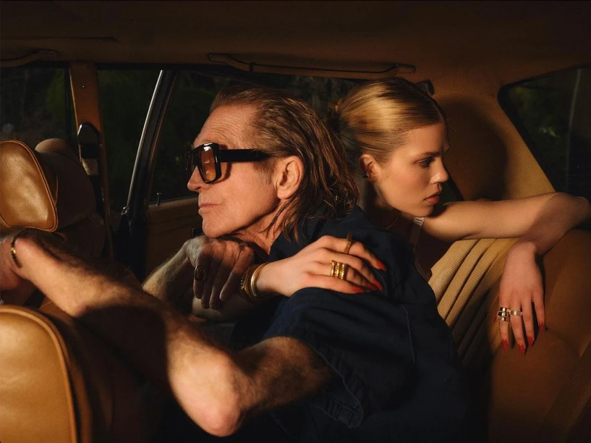 An older man with black sunglasses is posing in the back of a car with a young blond woman in a fashion editorial shot.