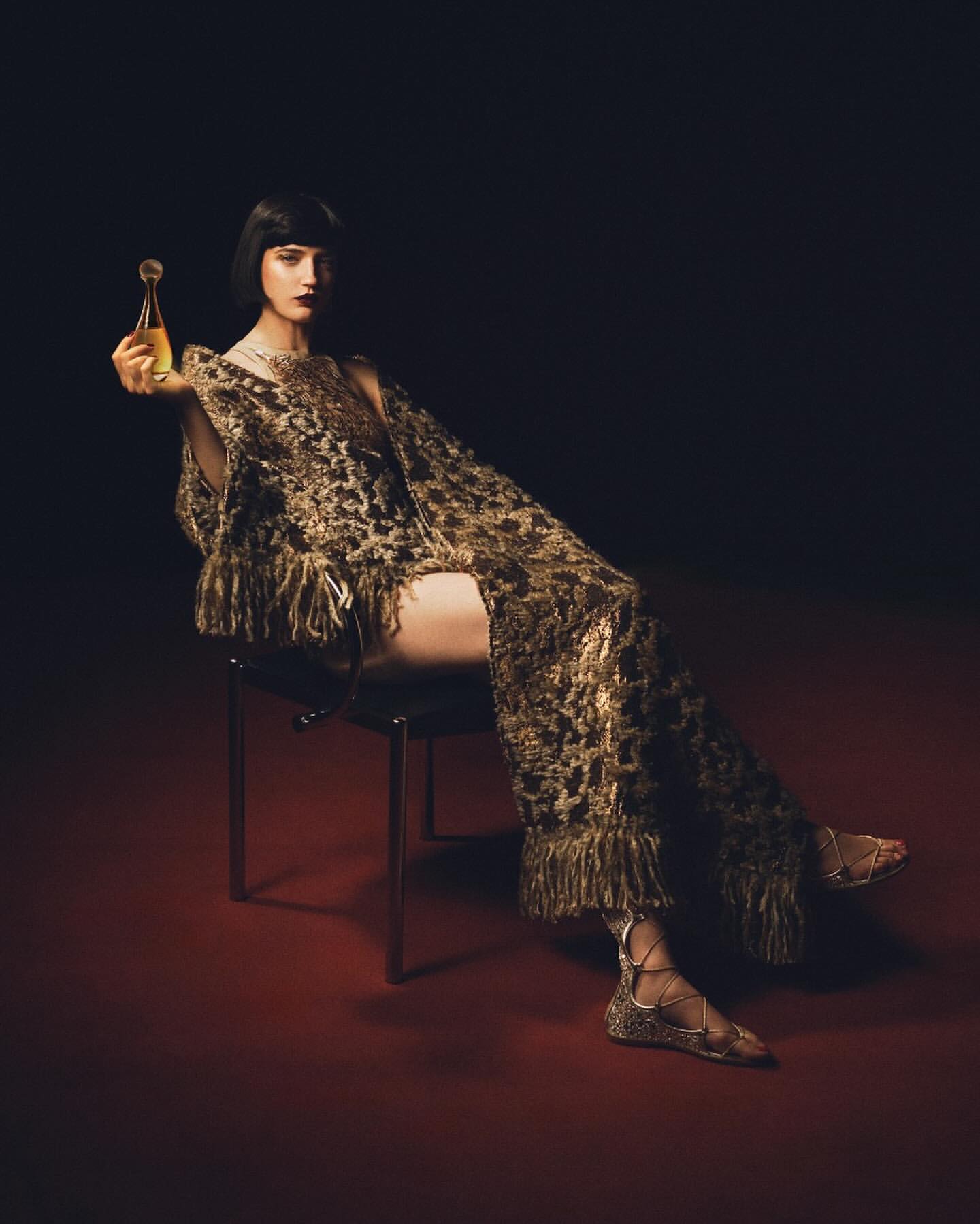 Brunette woman sits on chair in gold fringed outfit, holding gold perfume bottle against dark backdrop with red floor, studio lighting.