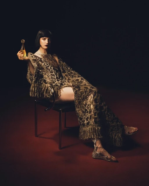 Brunette woman sits on chair in gold fringed outfit, holding gold perfume bottle against dark backdrop with red floor, studio lighting.