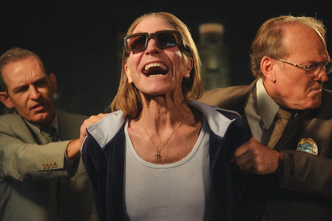 Middle-aged woman in sunglasses is escorted by two men in suits, seemingly against her will; she is yelling with her mouth open.
