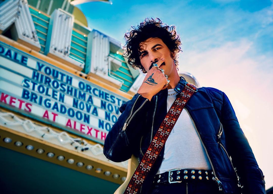 Adult male musician poses against the backdrop of a vintage theater. He is holding a cigarette. The theater sign advertises shows.