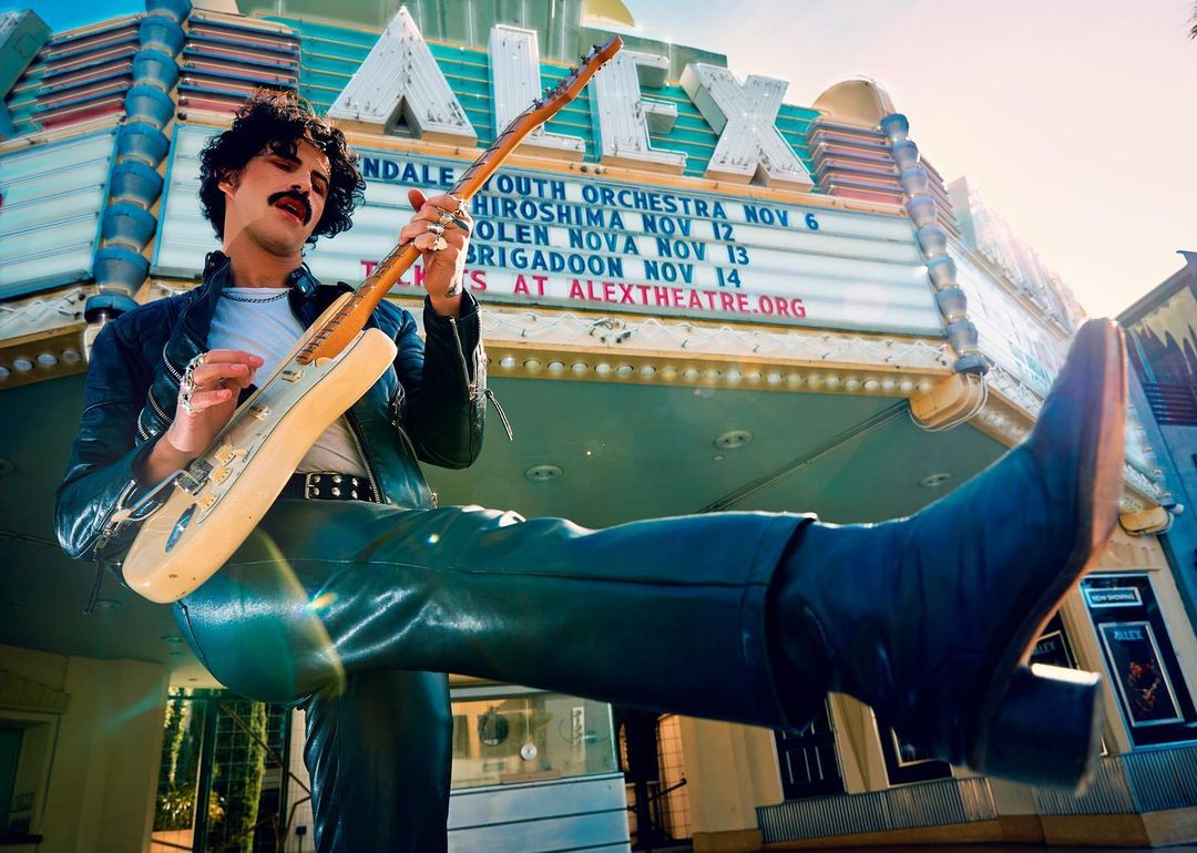 A musician with an electric guitar poses in front of the Alex Theatre, wearing a black leather jacket, black pants, and black boots.