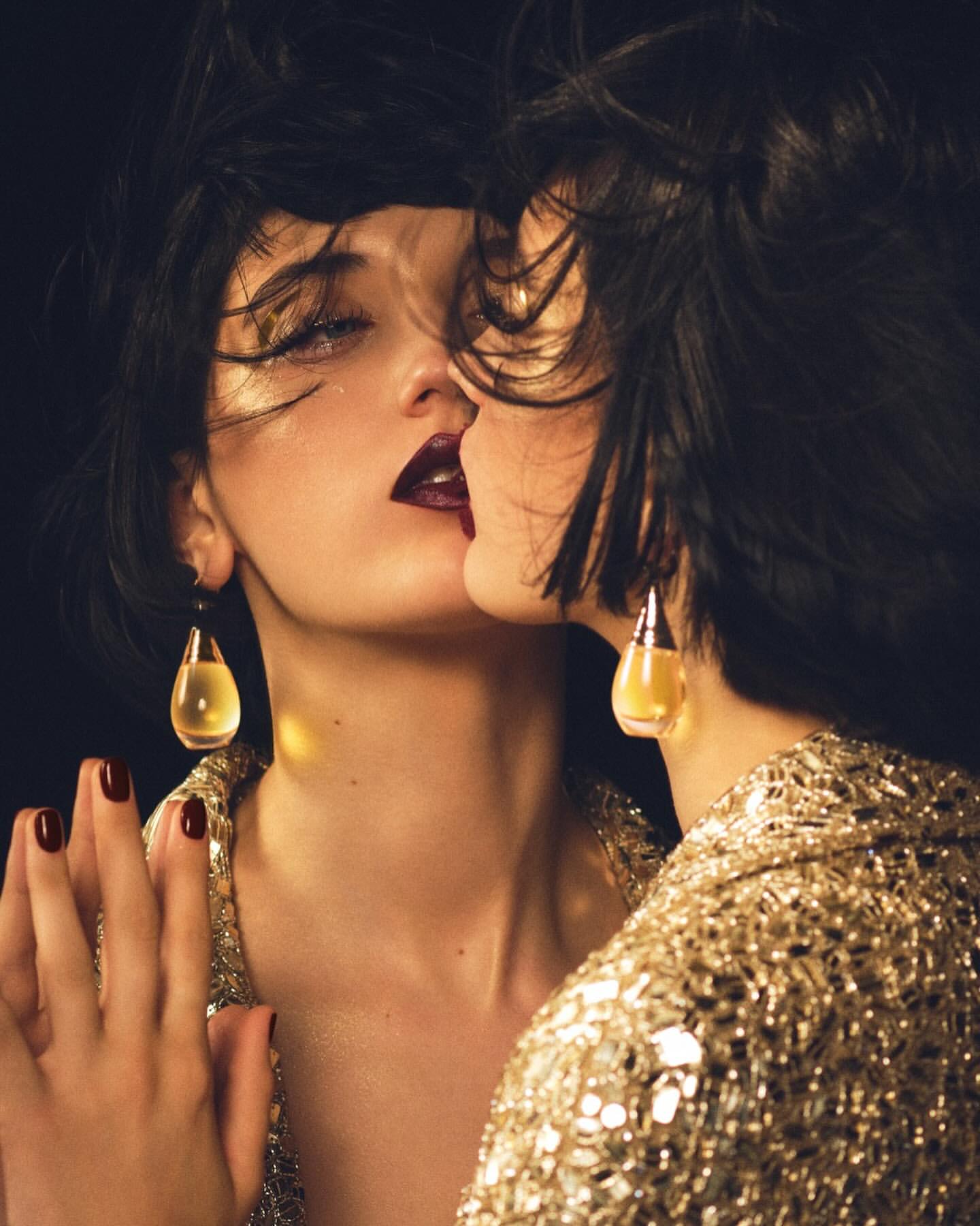 Close-up portrait of a woman with dark hair and dark lipstick reflected in a mirror, wearing a gold sequin jacket and teardrop earrings.