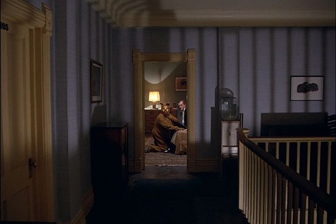 Interior view through doorway reveals a seated person with animal mask, and another person standing in the background near a lamp.