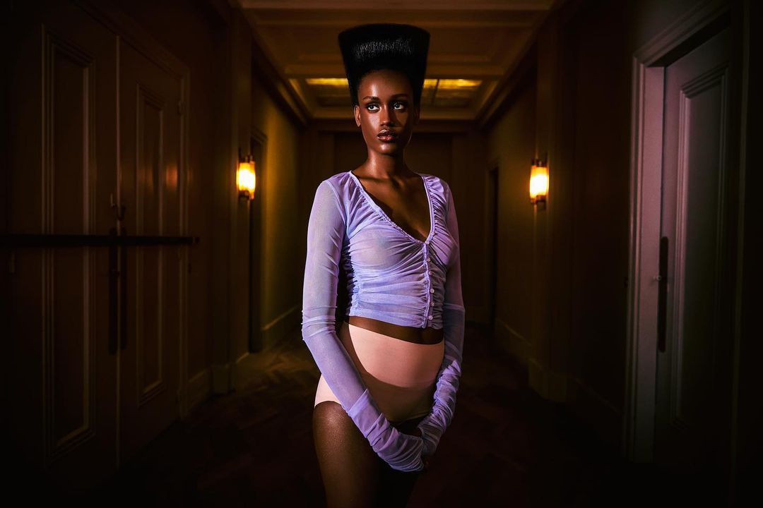 Portrait of an African American woman in a sheer top and briefs, posing in a hallway with warm sconce lighting and neutral wall tones.
