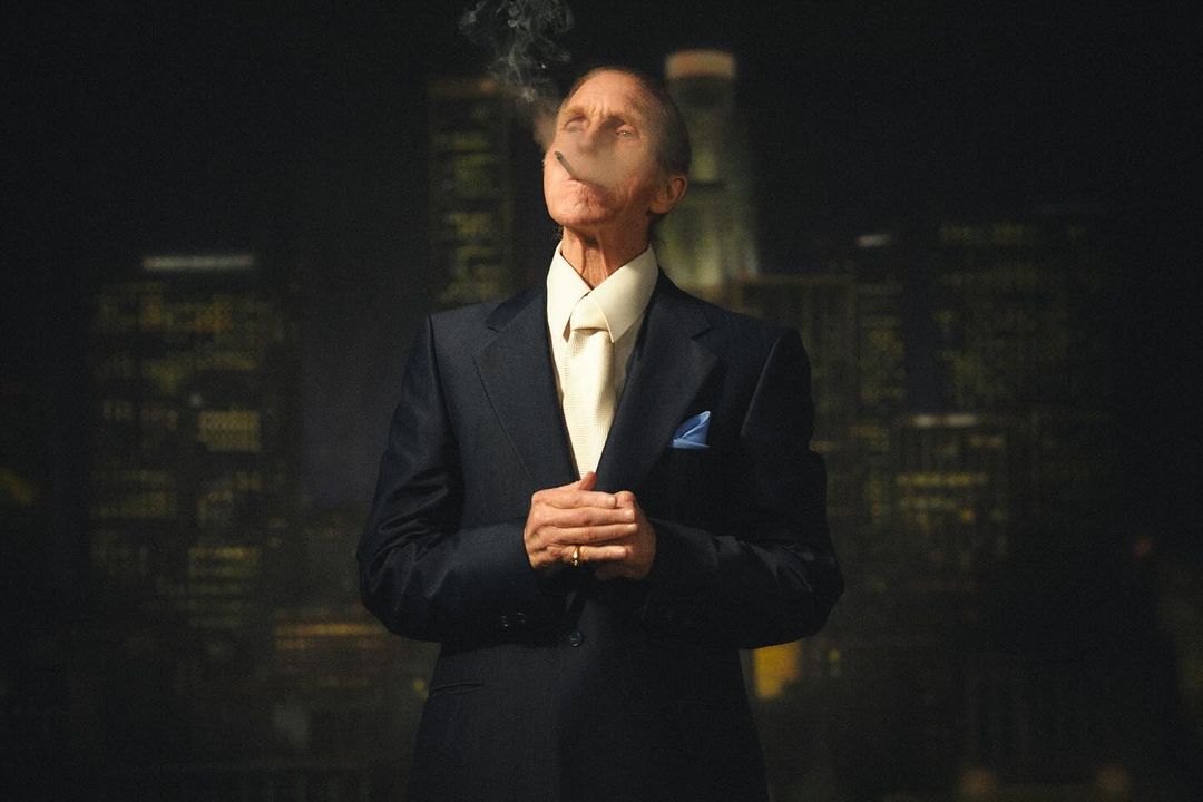 Elderly man in suit smoking, against a backdrop of city lights, creates a noir atmosphere with dark colors and low-angle shot.