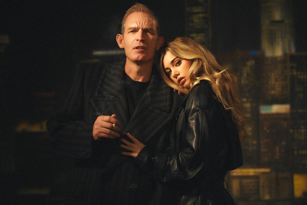 Indoor portrait of an adult man and adult woman in dark clothes, embracing in front of an abstract city backdrop with warm lighting.