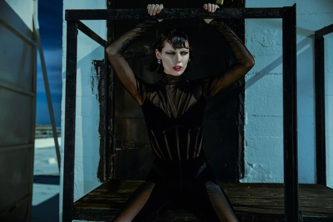 A woman in a black outfit poses holding onto metal bars, looking at the camera with a neutral expression in an indoor setting.