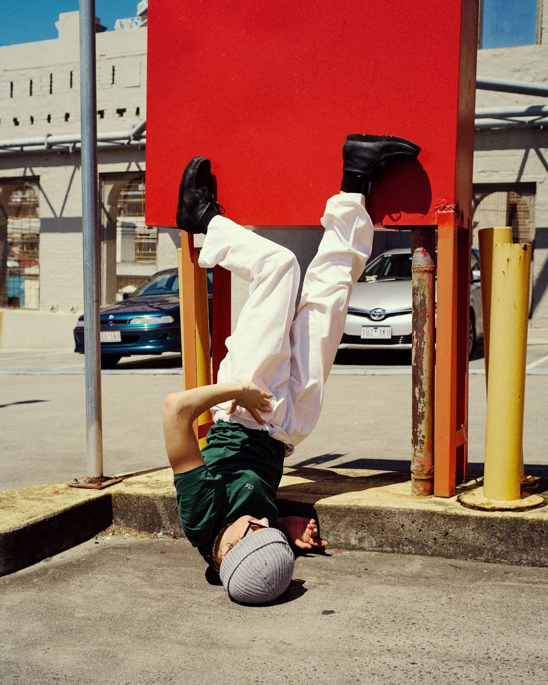 Young adult male in beanie, green shirt, and white pants performs a headstand against a red structure in an urban setting with cars.