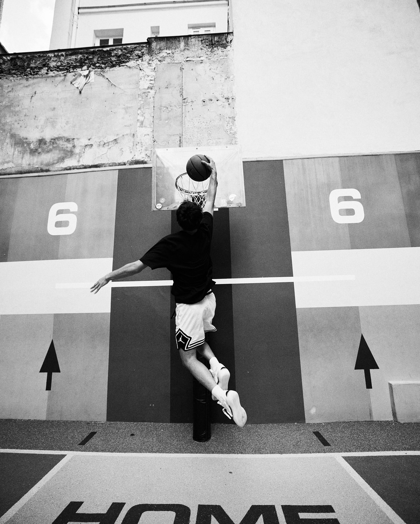 Black and white shot of a man playing basketball, mid-air, about to dunk the ball into the hoop outdoors on a painted court.