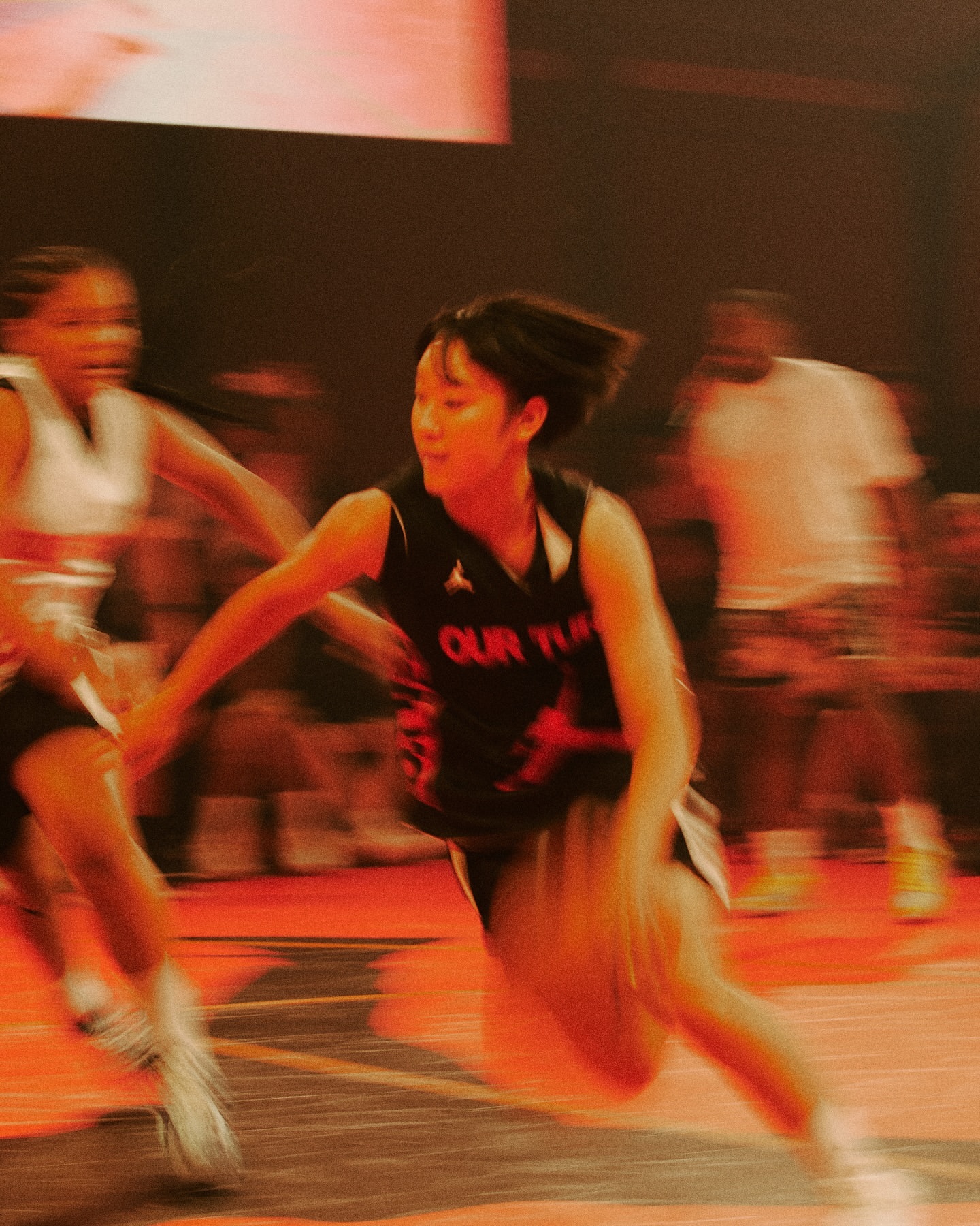 Female basketball player dribbling a ball during a basketball game with spectators, sports court with painted lines, orange-toned lighting.